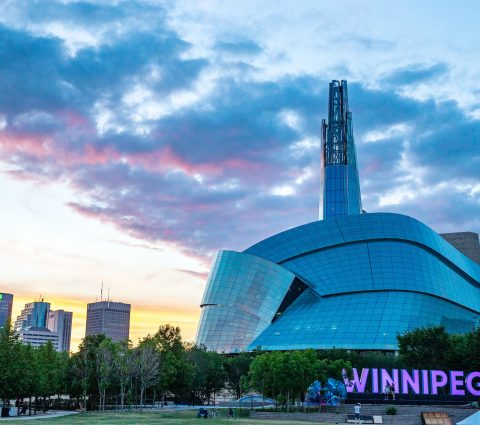Winnipeg sign lit in purple in front of the Canadian Museum for Human Rights at sunset.