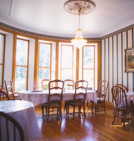 A bright dining room with many windows and hardwood floors, housing four tables covered in white table clothes and chairs.